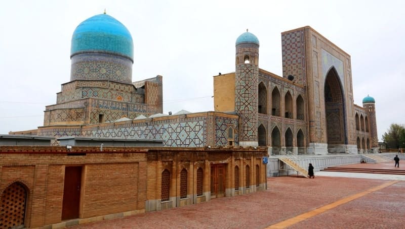 Blue dome and part of the facade of the Tillya-Kori Madrasah. The Tillya-Kori Madrasah (meaning "covered with gold") is part of the architectural ensemble of Registan Square.  A distinctive feature of this madrasah is the mosque located in its western part and crowned with a magnificent glass dome, visible in the photo.  The building is decorated with intricate blue tiles, typical of Samarkand, and elements covered in gold leaf.
